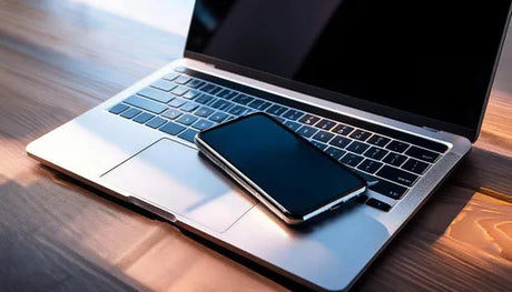 Black smartphone resting on a silver laptop, both placed on a wooden surface, showcasing a modern and sleek workspace.