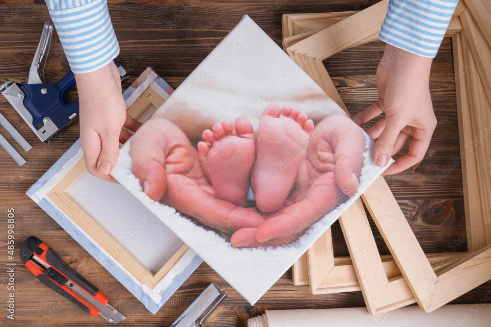 A person holds a canvas print of baby feet surrounded by hands, with framing tools and wood pieces scattered on a wooden surface.