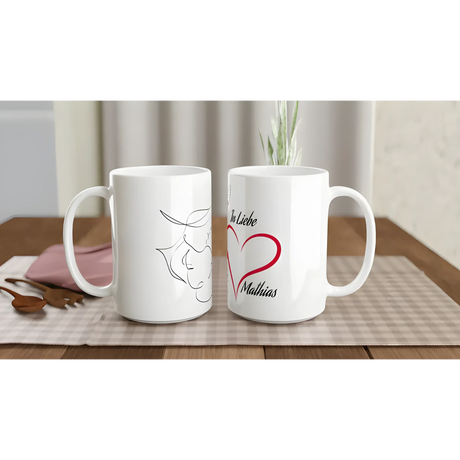 Two white ceramic mugs sit on a plaid tablecloth. One features a minimalist line drawing of a bull, while the other displays a red heart and the text "In Liebe Matthias."