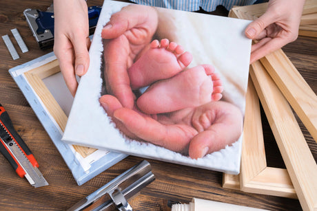 A hand holds a canvas print featuring baby feet resting in a parent's hands, surrounded by wooden framing materials on a wooden table.