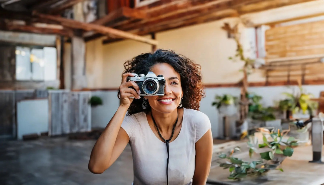 Smiling woman with curly hair holds a vintage camera in a bright, rustic setting with green plants and wooden décor.