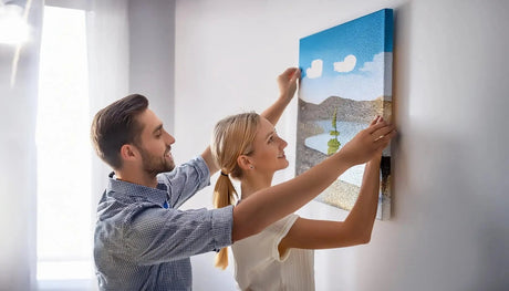 A smiling couple hangs a colorful canvas painting featuring a serene landscape on a light-colored wall in a bright room.