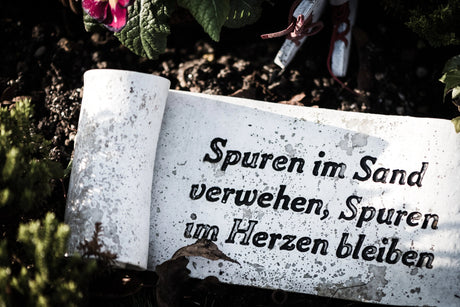 A weathered stone plaque with the inscription "Spuren im Sand verwehen, Spuren im Herzen bleiben" surrounded by green foliage and flowers.